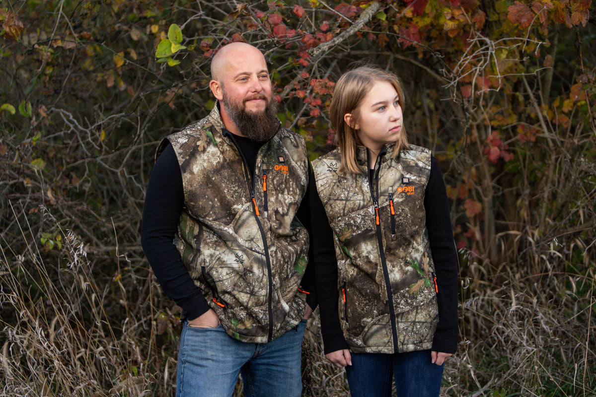 Father and daughter standing together in autumn brush, both wearing matching Gobi Heat Colorado heated vests in Realtree camo over black long-sleeve shirts with jeans.
