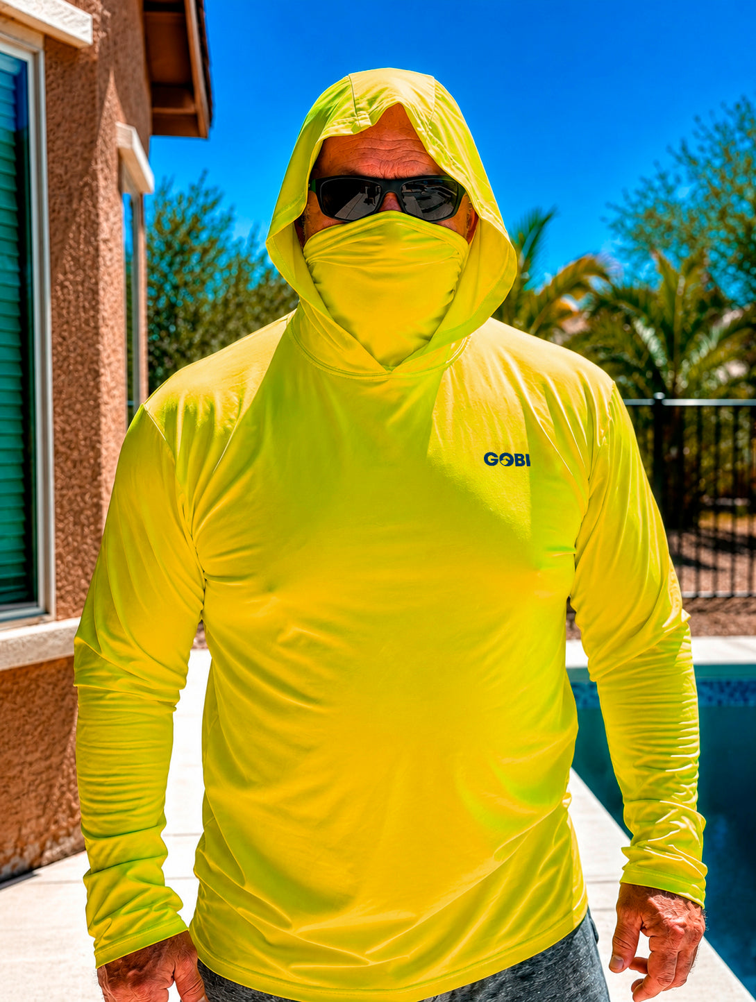 Man standing poolside in a hi-vis yellow Gobi Helio UPF Sun Hoodie with hood and face cover up, wearing sunglasses with a pool and palm trees behind him.