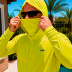 Close-up of a man pulling up the built-in face cover on a hi-vis yellow GOBI Helio UPF Sun Hoodie, wearing sunglasses outdoors with palm trees in the background.