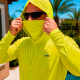 Close-up of a man pulling up the built-in face cover on a hi-vis yellow GOBI Helio UPF Sun Hoodie, wearing sunglasses outdoors with palm trees in the background.